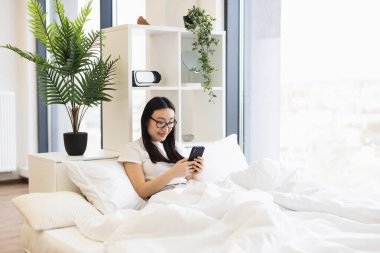 Genç Asyalı kadın akıllı telefon kullanarak yatakta dinleniyor. Doğal ışık ve yeşillikle modern yatak odası rahatlatıcı bir atmosfer yaratır..