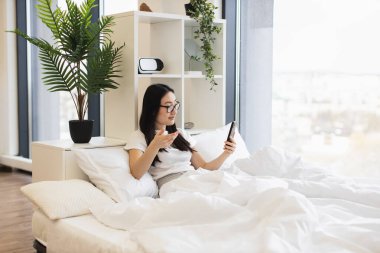 20 'li yaşlarda Asyalı bir kadın yatakta otururken online video görüşmesi için akıllı telefon kullanıyor. İç mekan bitkileri ve modern dekorla rahat bir atmosfer..