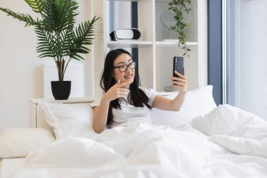 20 'li yaşlarda Asyalı bir kadın yatakta otururken online video görüşmesi için akıllı telefon kullanıyor. İç mekan bitkileri ve modern dekorla rahat bir atmosfer..