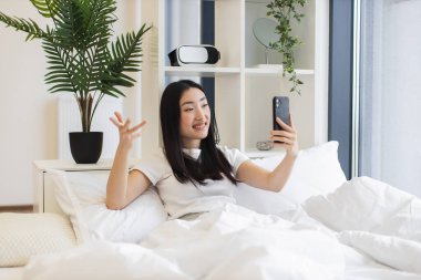 20 'li yaşlarda Asyalı bir kadın yatakta otururken online video görüşmesi için akıllı telefon kullanıyor. İç mekan bitkileri ve modern dekorla rahat bir atmosfer..