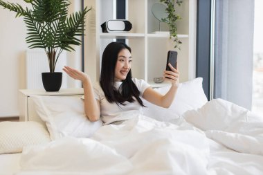 20 'li yaşlarda Asyalı bir kadın yatakta otururken online video görüşmesi için akıllı telefon kullanıyor. İç mekan bitkileri ve modern dekorla rahat bir atmosfer..