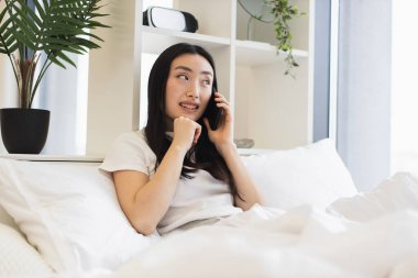 Yataktaki rahat Asyalı kadın telefonla konuşuyor. Genç yetişkinler rahat evlerinde sohbetin tadını çıkarıyorlar. Parlak oda, sakin atmosferi arttırır..