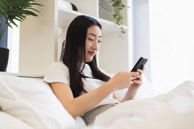Sosyal medya ya da mesajlaşma için akıllı telefon kullanan Asyalı bir kadın. Parlak ve rahat modern yatak odası, rahatlık ve rahatlık..
