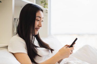 Asyalı bir kadın yatakta dinlenirken mesajları kontrol ediyor ya da sosyal medyaya akıllı telefondan bakıyor. Gündelik ve barışçıl atmosfer modern teknoloji ve iletişimi vurguluyor.