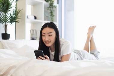 Genç Asyalı kadın rahat, parlak bir yatak odasında yatarken akıllı telefon kullanıyor. Boş zamanların tadını çıkarıyorum, internetten iletişim kuruyorum. Doğal ışık ile rahatlatıcı ve rahat ev ortamı.
