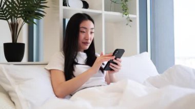 Genç Asyalı kadın akıllı telefon kullanarak yatakta dinleniyor. Doğal ışık ve yeşillikle modern yatak odası rahatlatıcı bir atmosfer yaratır..