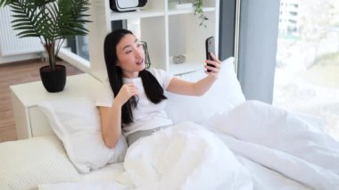 20 'li yaşlarda Asyalı bir kadın yatakta otururken online video görüşmesi için akıllı telefon kullanıyor. İç mekan bitkileri ve modern dekorla rahat bir atmosfer..