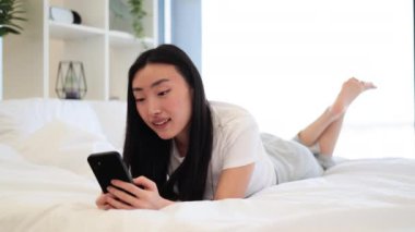 Genç Asyalı kadın akıllı telefon kullanarak yatakta uzanıyor, sosyal medya ve teknolojiden zevk alıyor. Parlak, huzurlu bir ortam, modern dekorlar, rahatlık ve rahatlık. Günlük ev ortamı.