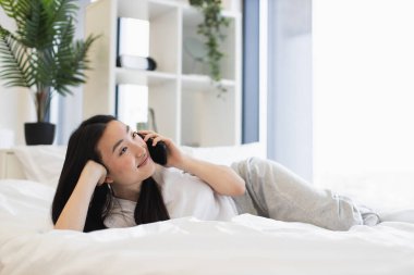 Asyalı genç bir kadın, aydınlık modern bir odada yatakta uzanırken telefonla konuşuyor, rahatlama ve neşe ifade ediyor..