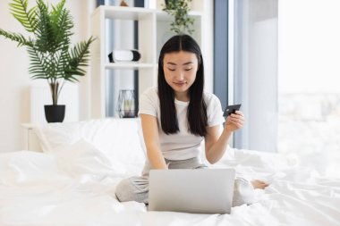 Genç Asyalı kadın yatağında otururken bilgisayardan alışveriş yapmak için kredi kartı kullanıyor. Modern teknoloji, kolaylık ve internet alışveriş kavramı.