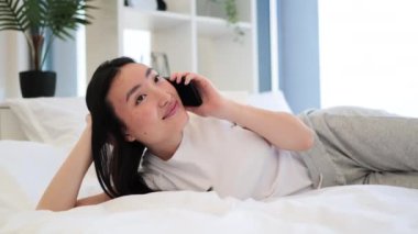 Genç Asyalı kadın telefonda konuşurken gülümsüyor. Parlak, modern bir yatak odası ortamında rahat ve memnun görünüyor..