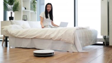 Genç Asyalı bir kadın modern yatak odasında robot elektrik süpürgesini kontrol etmek için uzaktan kumanda kullanıyor. Yatak konforundan temizlik teknolojisini yönetirken neşeli ifade.