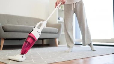 Genç Asyalı kadın, oturma odasının halısını elektrikli süpürgeyle süpürüyor. Günlük ev işlerini ve çağdaş yaşam tarzını vurgulayan temiz ve düzenli bir ev ortamı.