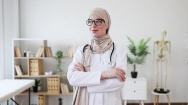 Başörtüsü ve gözlük takan genç bir müslüman kadın doktor portresi, özgüven ve profesyonellik yayıyor. Tıp alanında çeşitliliği, sağlık hizmetlerini ve yetkilendirmeyi temsil eder.