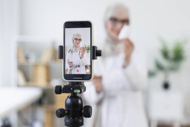 Türbanlı Müslüman bir kadın video kaydederken ilaç şişelerini gösteriyor. Genç yetişkin bir kadın, sağlık hizmetleri konusunda eğitici bir içerik yaratıyor. Profesyonel ortam çevrimiçi izleyicilerin ilgisini çekmeyi öneriyor.