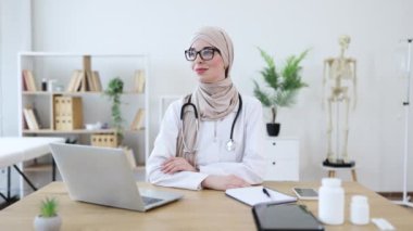20 'li yaşlarda, tesettürlü, ofis masasında oturan Orta Doğulu bir kadın doktor. Laptop, notlar ve steteskopla tıbbi ayar. Profesyonel ve kendine güvenen atmosfer sağlık ortamını betimliyor.