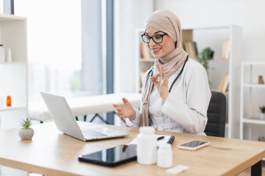 Genç Müslüman kadın doktor, modern ofiste dizüstü bilgisayar kullanarak hastasıyla görüntülü görüşme yapıyor. Orta Doğulu kadın sağlık iletişim teknolojisini resmediyor.