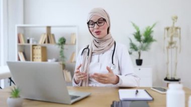 Orta Doğulu kadın doktor tesettüre girip dizüstü bilgisayarla konsültasyon yapıyor. Teletıp, uzaktan sağlık hizmetlerine odaklan. Modern ve profesyonel ofis ayarları çevreyi geliştirir.