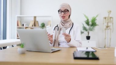 Genç Müslüman kadın doktor, video görüşmesi sırasında hap tutuyor. Başörtüsü ve gözlük takarak sanal sağlık hizmetine girmiş. Orta Doğulu kadın modern ofiste çevrimiçi tıbbi danışmanlık yapıyor.