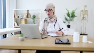 Genç Müslüman kadın doktor online danışmanlık için dizüstü bilgisayar kullanıyor, notlar yazıyor. Modern ofiste çalışan bir tıp uzmanı. Başörtüsü içinde kendine güvenen yetişkin bir kadın uzaktan sağlık hizmeti sunuyor..