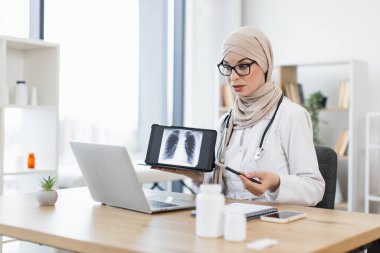 Orta Doğulu tesettürlü kadın doktor video çağrısı sırasında akciğer röntgeniyle birlikte tablet tutuyor. Görüntü teletıp, tıbbi konsültasyon ve hasta bakımını betimliyor.