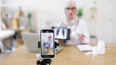 Genç Müslüman kadın doktor cep telefonuyla dersleri kaydediyor. Ciğerlerinin röntgen sonuçlarını açıklıyor. Parlak bir ofiste tıbbi ekipman kullanıyor, eğitim ve sağlık hizmetleri iletişimine odaklanıyor..