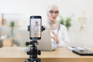Genç Müslüman kadın blogu için telefonda video dersi kaydediyor. Modern çalışma alanında dizüstü bilgisayar ve tripod.