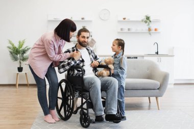 Tekerlekli sandalyede babası olan beyaz bir aile anne ve küçük kızıyla sıcak etkileşim içinde. Modern mutfak misafirperver bir ortam yaratır. Aile sevgisi, destek ve maluliyet bilincini yakalar.