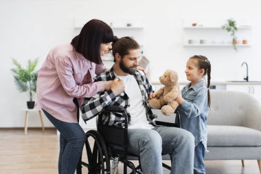 Tekerlekli sandalyede babası olan beyaz bir aile anne ve küçük kızıyla sıcak etkileşim içinde. Modern mutfak misafirperver bir ortam yaratır. Aile sevgisi, destek ve maluliyet bilincini yakalar.