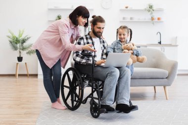 Modern mutfakta beyaz bir aile, anne tekerlekli sandalyede sakat bir baba, oyuncak ayıyla genç bir kız. Sıcak aile anları sevgi ve destek gösteriyor..