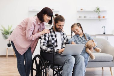 Tekerlekli sandalyedeki babası, annesi ve küçük kızı modern mutfakta eğlenen beyaz bir aile. Hepsi dizüstü bilgisayara odaklanırken kızı oyuncak ayıyı tutuyor..