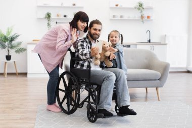 Modern mutfak ortamında babası tekerlekli sandalyede, annesi ve küçük kızı olan beyaz bir ailenin portresi. Mutlu an, ailenin birlikteliğini, sevgisini ve uyum sağlayabilirliğini gösteriyor..