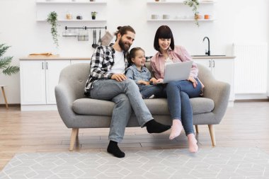 Annesi, babası ve genç kızıyla birlikte beyaz bir aile kanepede oturmuş dizüstü bilgisayar izliyor. Modern oturma odasında gündelik, keyifli bir ortam. Teknoloji ve bağlanma ile mutlu, rahat bir atmosfer.
