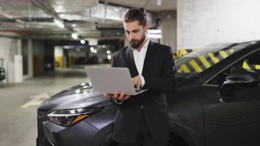 Takım elbiseli beyaz bir erkek yeraltı otoparkında dizüstü bilgisayar kullanıyor. İş kurmak profesyonelliği yansıtır ve teknolojiye odaklanır. Kentsel çevre kurumsal atmosferi güçlendirir.