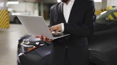 Otoparkta dizüstü bilgisayarla çalışan beyaz işadamı daktilo üzerine odaklanmış takım elbise giyiyor. Teknoloji, iş hareketliliği ve modern çalışma ortamlarını temsil eder.