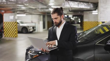 Takım elbiseli beyaz erkek işadamı yeraltı otoparkında dizüstü bilgisayar kullanıyor. Profesyonel ve odaklanmış olarak, kentsel ortamlarda modern teknoloji kullanımı ve çalışmaları temsil etmektedir..