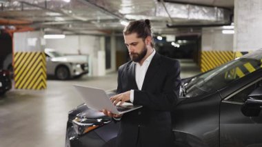 Takım elbiseli beyaz bir erkek işadamı elinde dizüstü bilgisayar tutarak şehrin yeraltı otoparkında baş parmağını kaldırıyor, arabanın yanında duruyor, iş yerinde başarı ve güven gösterisi yapıyor..