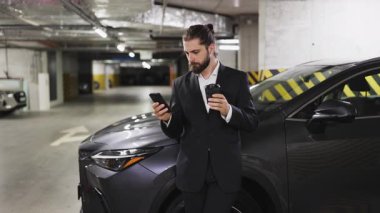 Kafkas 'lı genç iş adamı kahve tutuyor ve otoparkta telefon kullanıyor. Odaklanmış ve sıradan görünüyor, yeraltı garaj ortamında arabaya yaslanıyor..