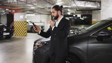 Takım elbiseli genç beyaz bir erkek yeraltı otoparkında arabasının yanındaki telefon görüşmesinde konuşuyor. Teknoloji ve modern çalışma tarzına odaklı iş iletişimi kavramı.