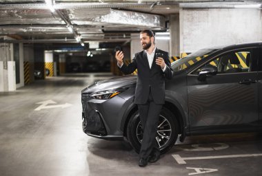 Takım elbiseli genç beyaz erkek garajda video görüşmesi için akıllı telefon kullanıyor, elinde sıcak içki, arabanın yanında..