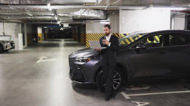 Genç beyaz erkek işadamı video görüşmesi için otoparkta dizüstü bilgisayar kullanıyor. Arabanın yanında gelişigüzel dikiliyorsun. Resmi giyinmiş, odaklanmış ve profesyonel görünüyor..