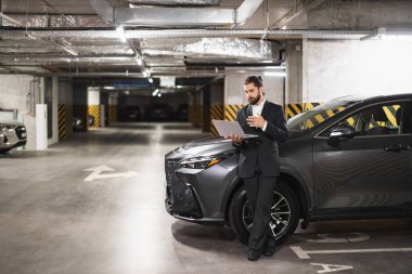 Genç beyaz erkek işadamı video görüşmesi için otoparkta dizüstü bilgisayar kullanıyor. Arabanın yanında gelişigüzel dikiliyorsun. Resmi giyinmiş, odaklanmış ve profesyonel görünüyor..