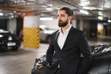 Yeraltı otoparkında takım elbiseli beyaz erkek işadamının portresi, güven ve profesyonellik yayıyor. Arka planda çok sayıda park edilmiş araba ve endüstriyel ışıklandırma yer alıyor.