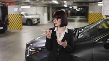 Genç, beyaz bir kadın modern bir otoparkta akıllı telefondan sesli mesaj kaydediyor. İş kıyafetleri, iletişim ve teknoloji kavramları ile profesyonel ortam.
