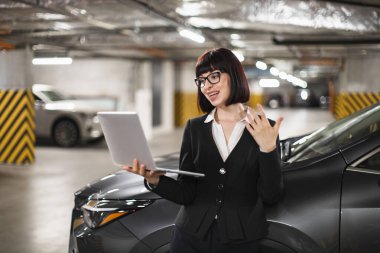 Beyaz bir iş kadını kapalı otoparkta dizüstü bilgisayarla arabaya yaslanmış online video görüşmesi yapıyor ve fikirlerini dile getiriyor. Uzaktan çalışan profesyonel giyimli genç bir kadın..