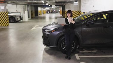 Akıllı giyimli beyaz kadın kapalı otoparkta bilgisayar kullanarak video çekiyor. Arka planda araçları olan profesyonel ortam, modern iş ortamını yansıtıyor.