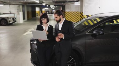 Beyaz adam ve kadın iş kıyafetleri içinde otoparkta laptop belgelerini gözden geçiriyorlar. Profesyonel işbirliği, takım çalışması atmosferi, kentsel ortam. İkisi de takım elbiseli, göreve odaklanmış..