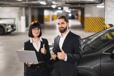 Beyaz iş kadını ve işadamı gülümsüyor, yeraltı otoparkında başparmak kaldırıyor. Laptoplu ve kahveli profesyonel kıyafet. Takım çalışmasını, başarıyı, şehir ofisine olan güveni gösterir.