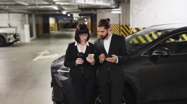 Beyaz erkek ve kadın iş kıyafetleri içinde akıllı telefonları park halindeki arabaların yanında kullanıyorlar. Bağlanabilirlik ve iletişim odaklı modern, profesyonel ortam.
