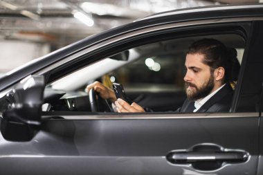 Takım elbiseli beyaz erkek, kapalı alanda park etmiş arabanın içinde akıllı telefon kullanıyor. Genç yetişkin, dikkati dağılmış sürüş konseptini vurgulayarak cihaza odaklanmış görünüyor..